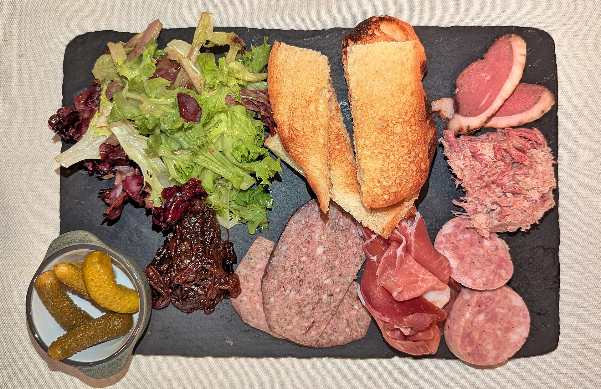 Charcuterie board with salami, prosciutto, crostini, and pickles on a slate board
