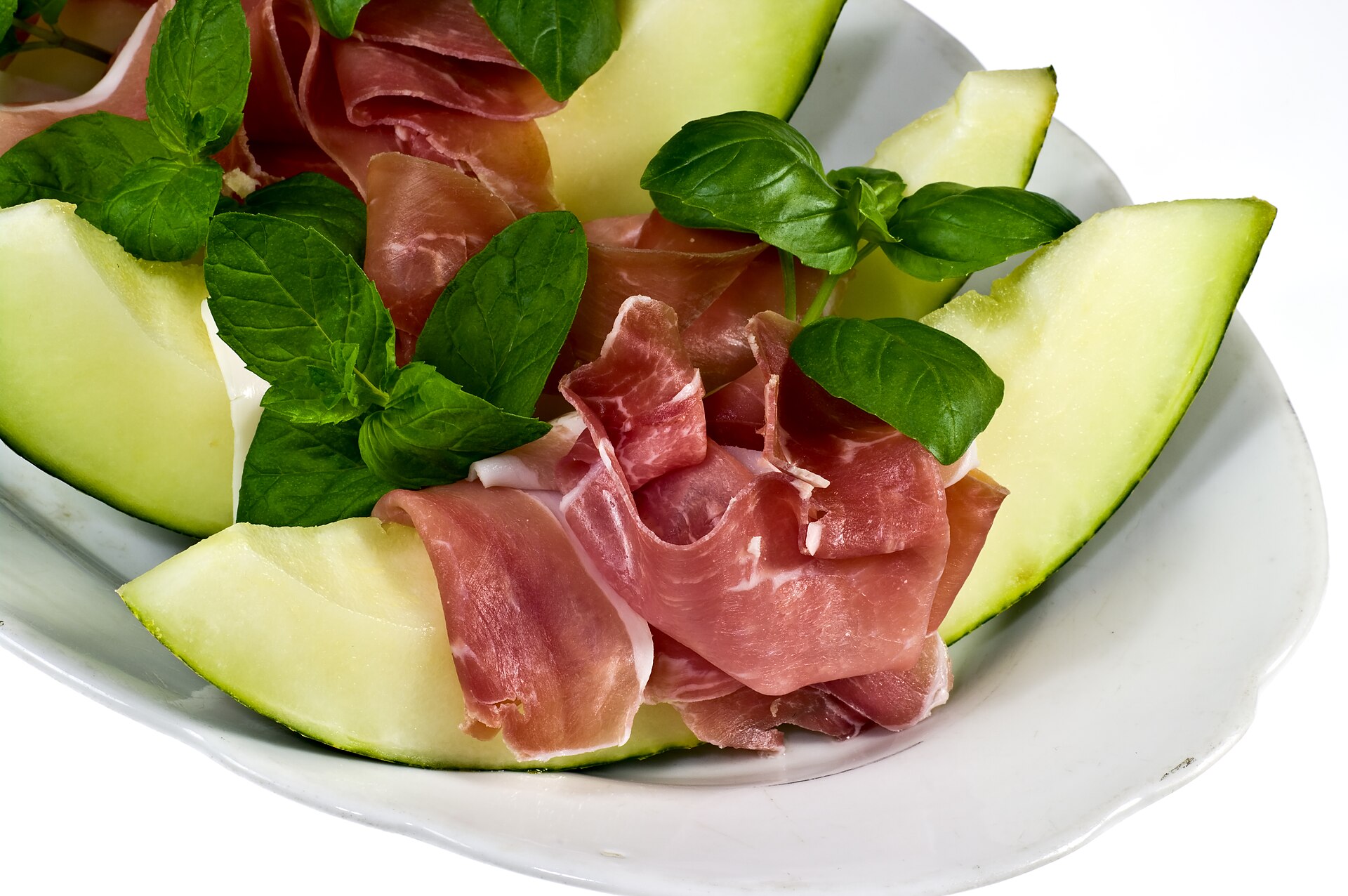 Prosciutto draped over melon wedges with basil on a white plate